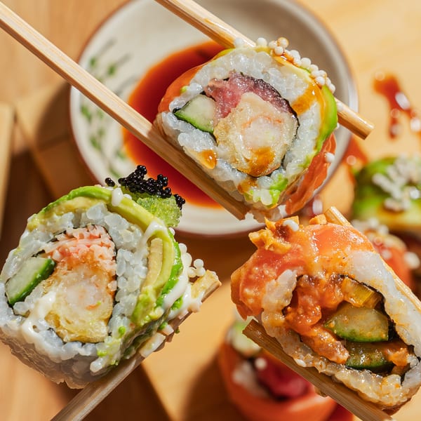 Three different sushi rolls, held by chopsticks, with dipping sauce in the background.