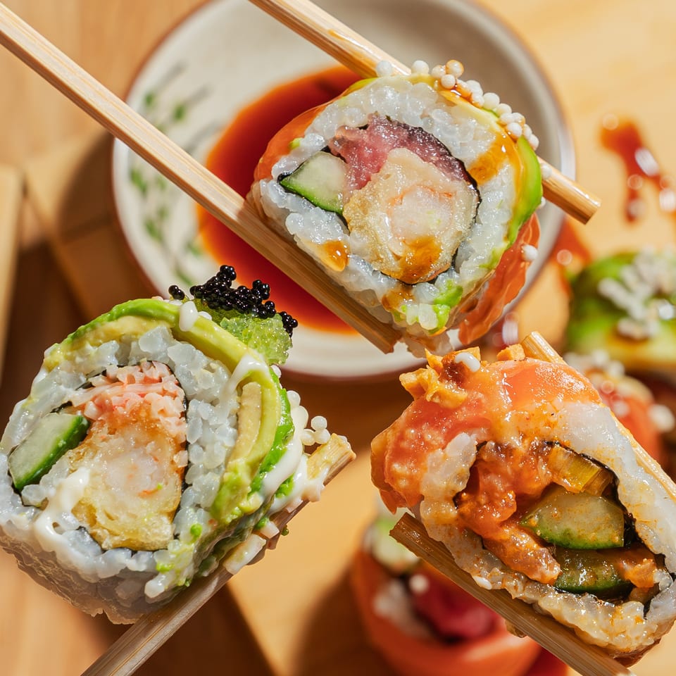 Three different sushi rolls, held by chopsticks, with dipping sauce in the background.