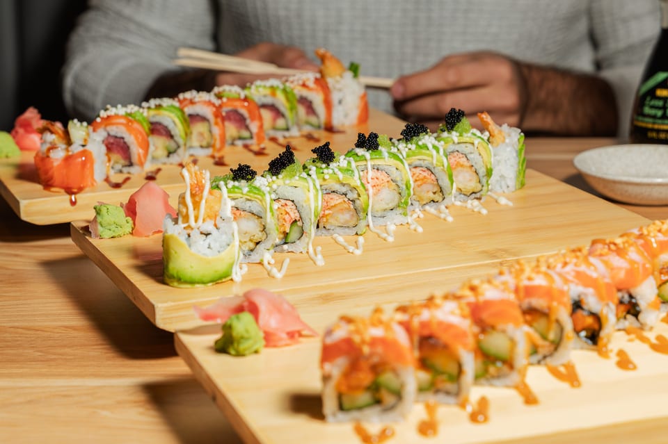 Three rows of sushi rolls with various toppings, wasabi, and ginger on wooden boards.